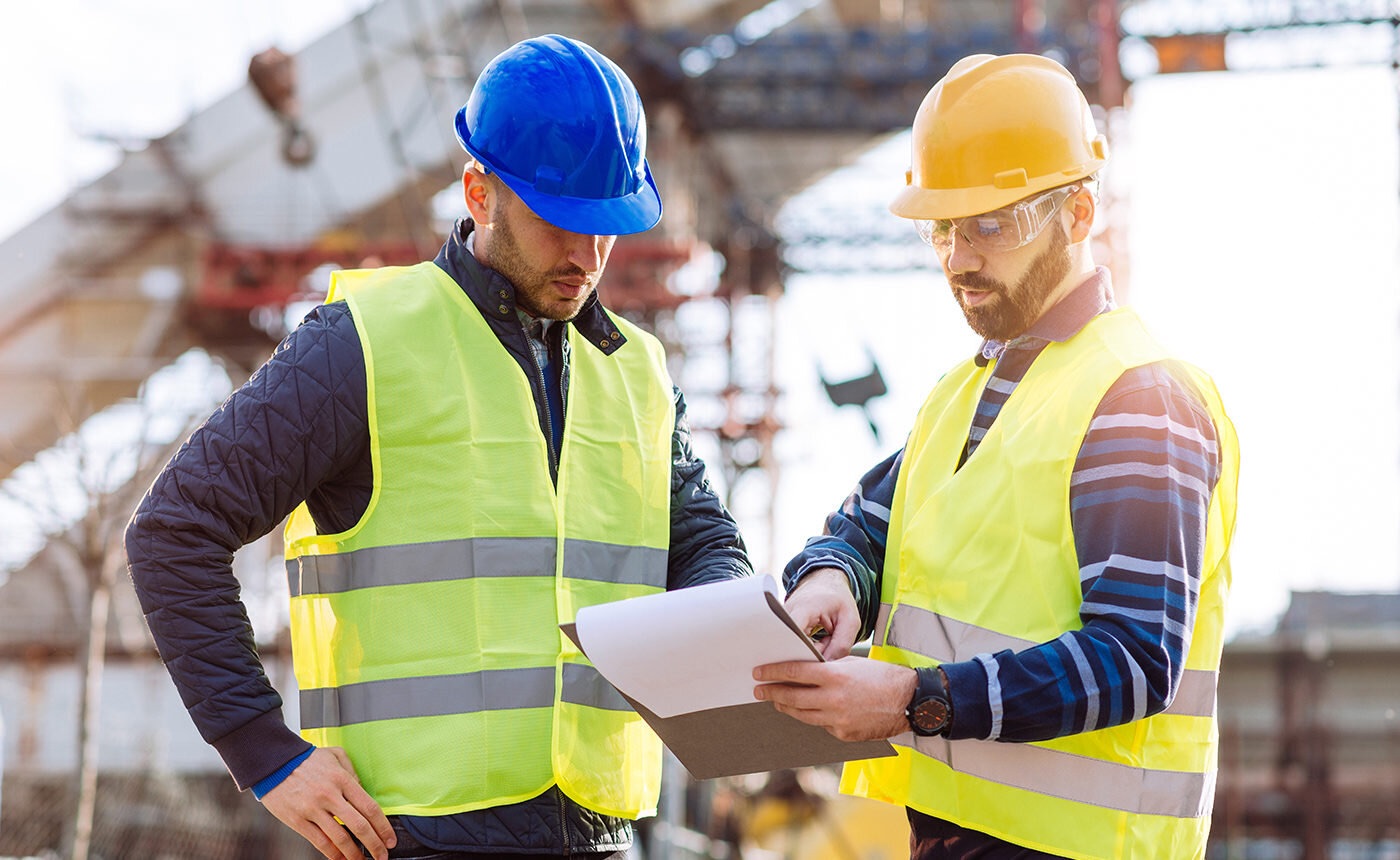 Two male technicians in hard hats and safety vests are reviewing documents at a construction site.